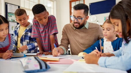 Young school teacher helping elementary students while writing in notebooks. Hispanic male teacher wearing spectacles and checking student notebooks after test while talking to them in classroom. Happy male teacher helping and checking works of pupil in classroom.
