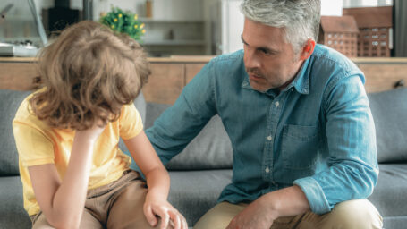 Sad little kid sitting on sofa at room, looking away and talking with mature father