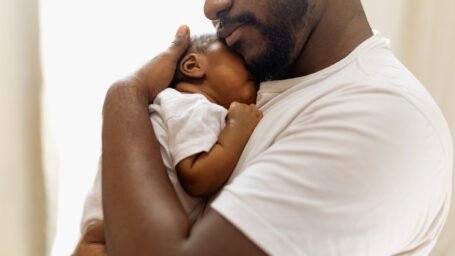 Childcare concept. Loving and caring African American father giving a hug to his newborn baby daughter at home, young black father spending time with his cute little daughter, in the living room, copy