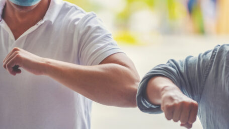 Men wearing surgical face masks practicing social distancing outdoors. They are doing an elbow bump greeting. Both are casually dressed.