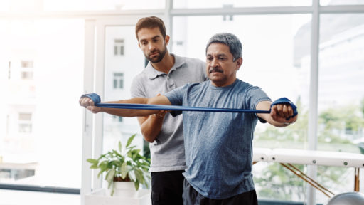 Image of a young male physiotherapist helping a mature male patient with movement exercises at a clinic.