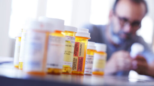A large group of prescription medication bottles sit on a table in front of a man who is holding and looking at one of the pill bottles out of focus in the background. The image is photographed with a very shallow depth of field with the focus being on the pill bottles in the foreground.