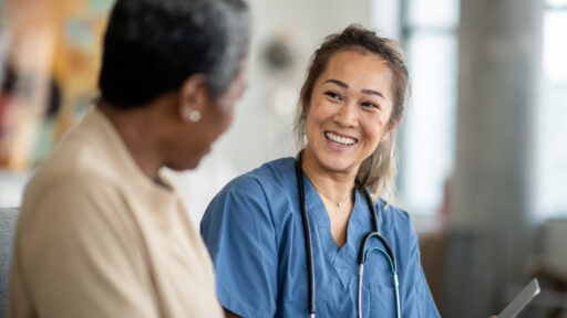 A female nurse of Asian decent, sits with a senior patient in the comfort of her own living room during a Homecare visit. She is wearing blue scrubs and has a tablet in her hands as she reviews some recent test results with her patient. The senior woman is dressed casually and is asking the nurse a question about the results.