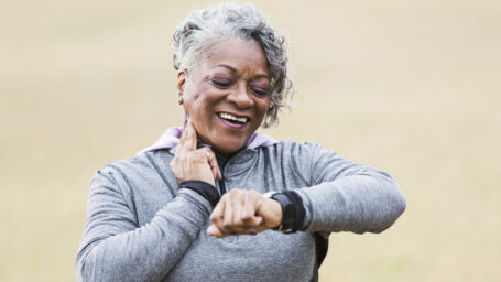 An African American senior woman, in her 60s, exercising outdoors in the park. She is checking her pulse to see if her exercise has increased her heart rate. She is smiling, with two fingers on her neck, looking at the watch on her wrist. She is wearing a gray sweatshirt.