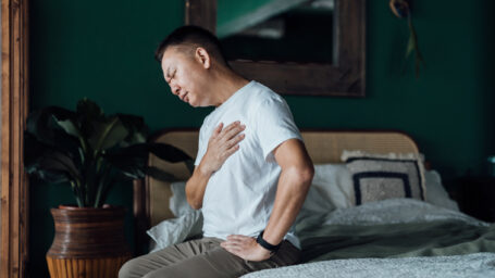 Senior Asian man with eyes closed holding his chest in discomfort, suffering from chest pain while sitting on bed at home. Elderly and health issues concept