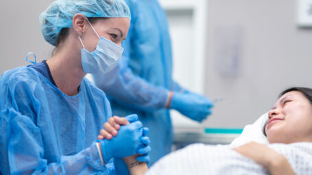 A female surgeon is with her team of surgeons. The multi-ethnic group is preparing to perform a cesarian section on a pregnant woman. The female doctor is sitting next to a patient who is lying on an operating table. The doctor is holding the patient's hand while providing comfort.