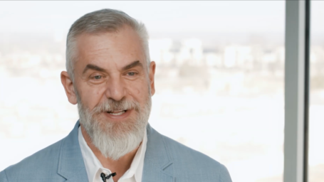 Dr. W. Cody Grammer in a light blue blazer and white shirt speaking on camera in a bright, modern medical office setting with large windows in the background.