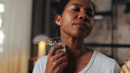 African American woman in pajamas sitting on bed, fanning herself and drinking ice water during menopause-related hot flashes. Heat relief, wellness, home comfort and self-care concept.