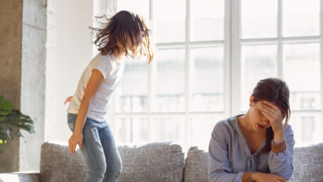 Upset mother having problem with noisy naughty daughter jumping on couch and screaming, demanding attention, frustrated mum tired of difficult child, child tantrum manipulation concept