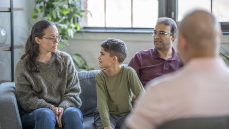 A family of three sit on a couch in front of a counselor and discuss mental health struggles in an urban office space.