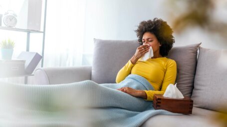 Sick young woman sitting on sofa blowing her nose at home in the sitting room. Photo of sneezing woman in paper tissue. Picture showing woman sneezing on tissue on couch in the living-room (Sick young woman sitting on sofa blowing her nose at home in